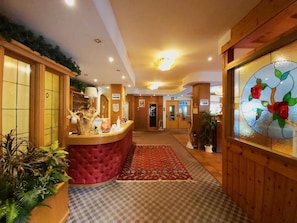 A reception area with a red cushioned counter, a checkered carpet, and a glass wall with a floral design. - Hotel Scoiattolo (Falcade)