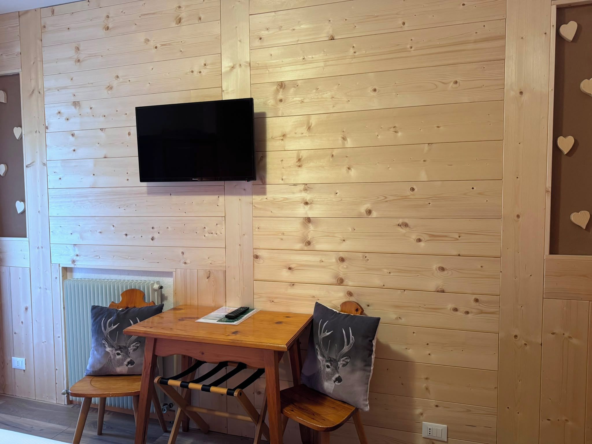 A wooden-paneled room with a flat-screen TV mounted on the wall, a wooden table with two chairs, and decorative heart-shaped cutouts.