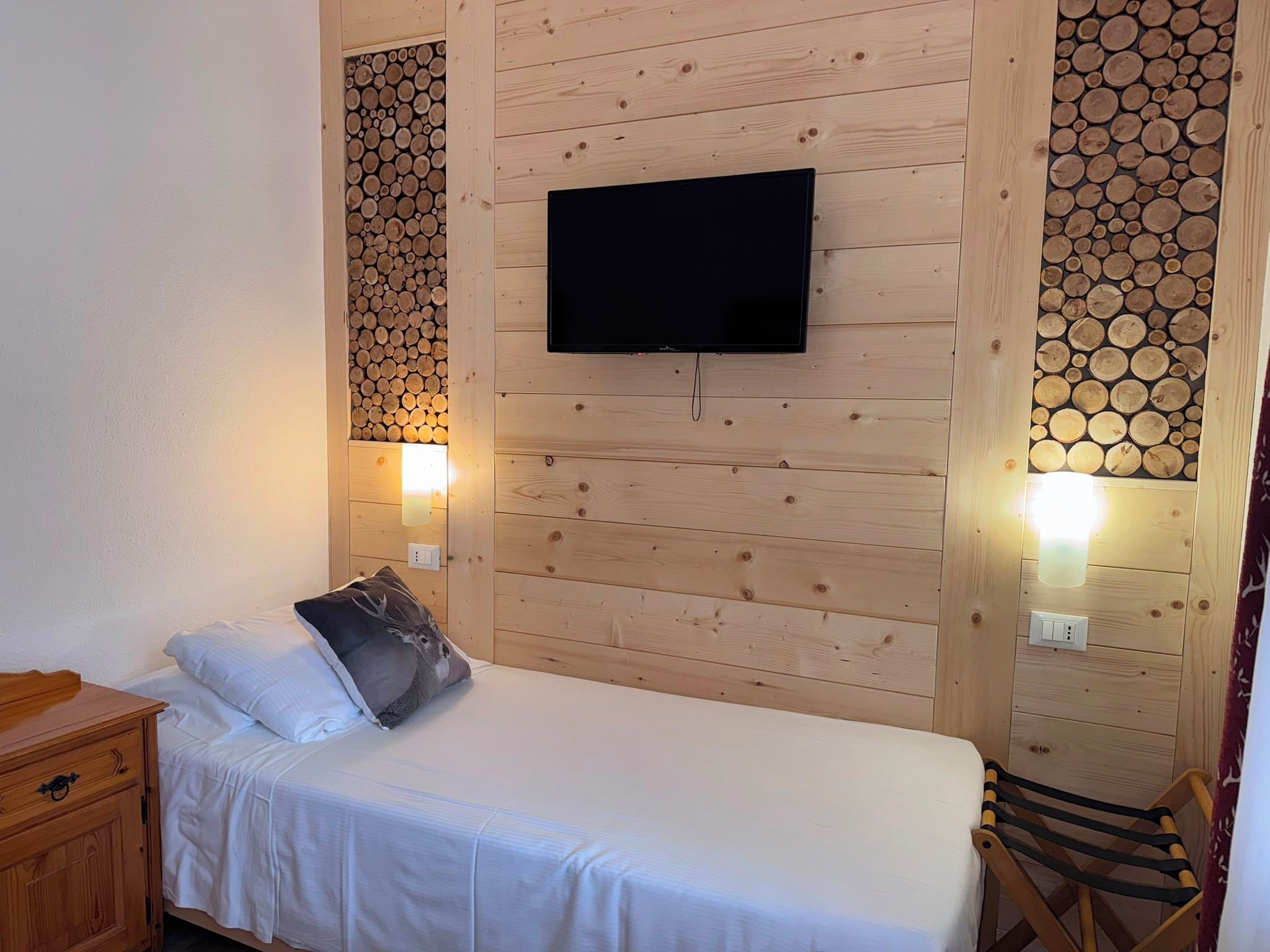 A single bed with a white mattress and a gray pillow, a flat-screen TV mounted on a wooden wall, and two wall-mounted lights.