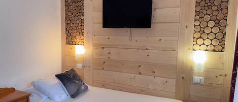 Een enkel bed met een wit matras en een grijs kussen, een flatscreen televisie aan een houten wand bevestigd, en twee wandlampen.