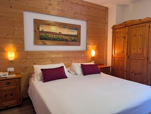 A hotel room with a large bed, wooden headboard, two bedside tables, a nightstand, a wardrobe, and a framed landscape painting on the wall. - Hotel Scoiattolo (Falcade)