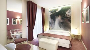 A modern hotel room with a large bed, a clear chair, a small table, and a large painting on the wall.