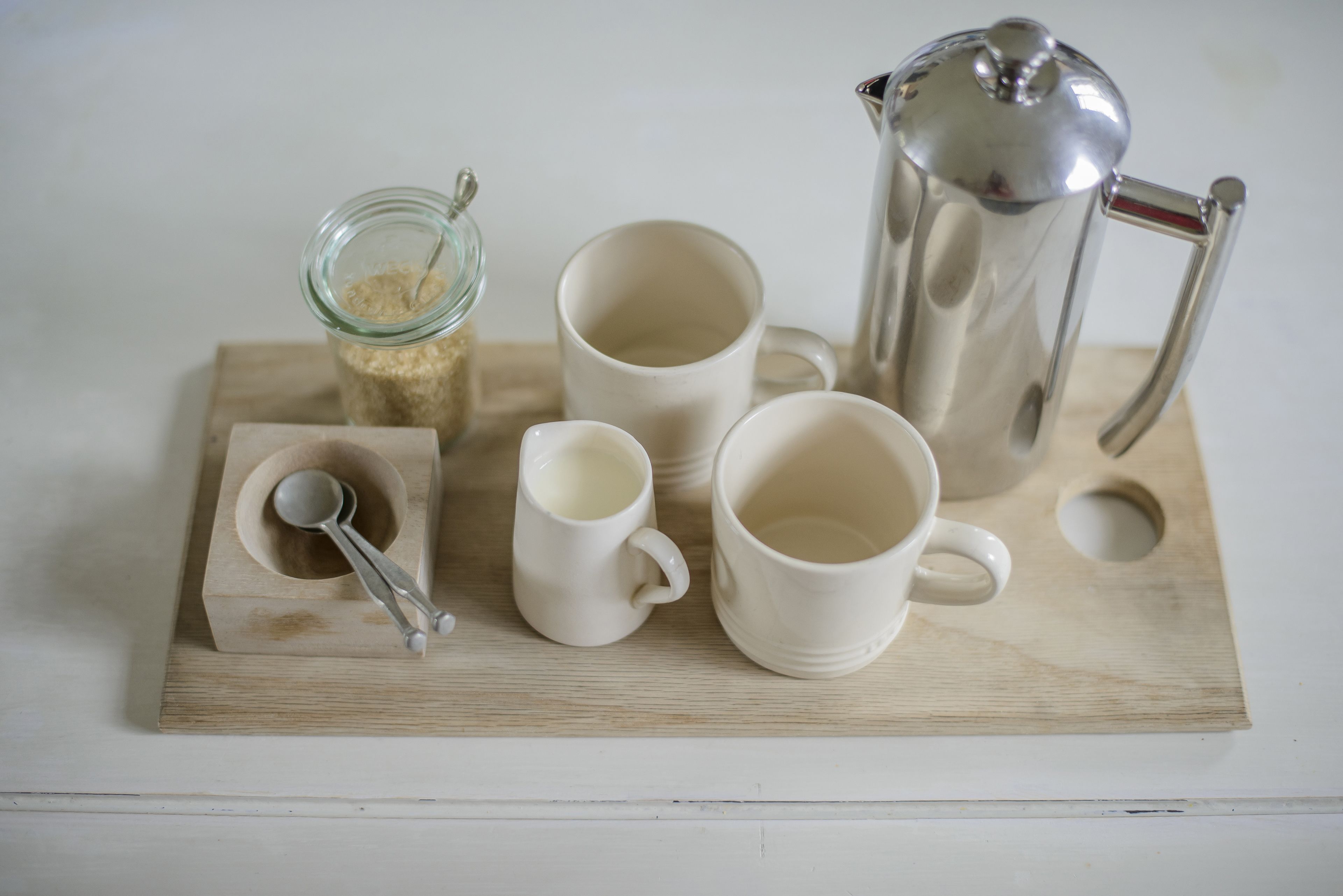 A wooden tray with a coffee maker, two cups, a spoon, and a jar of sugar.