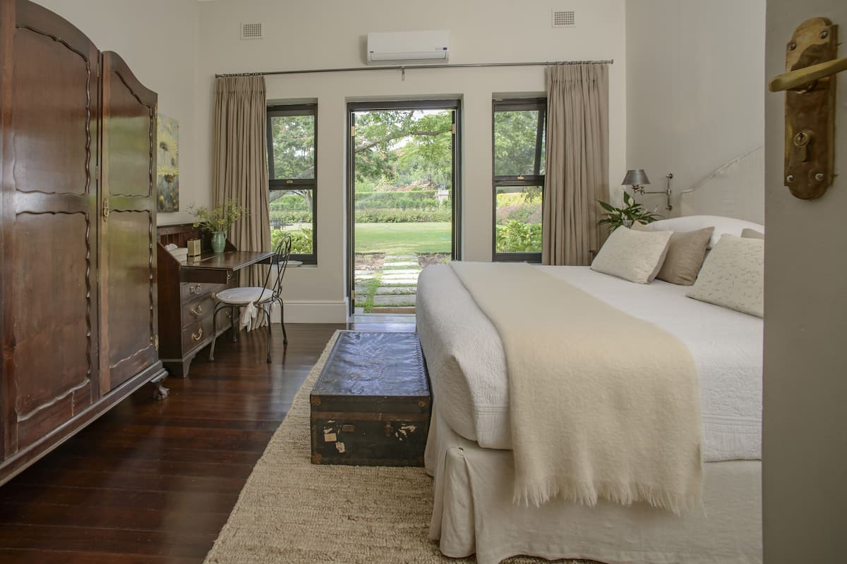 A bedroom with a large bed, a wooden wardrobe, a desk with a chair, and a view through a glass door to a garden.