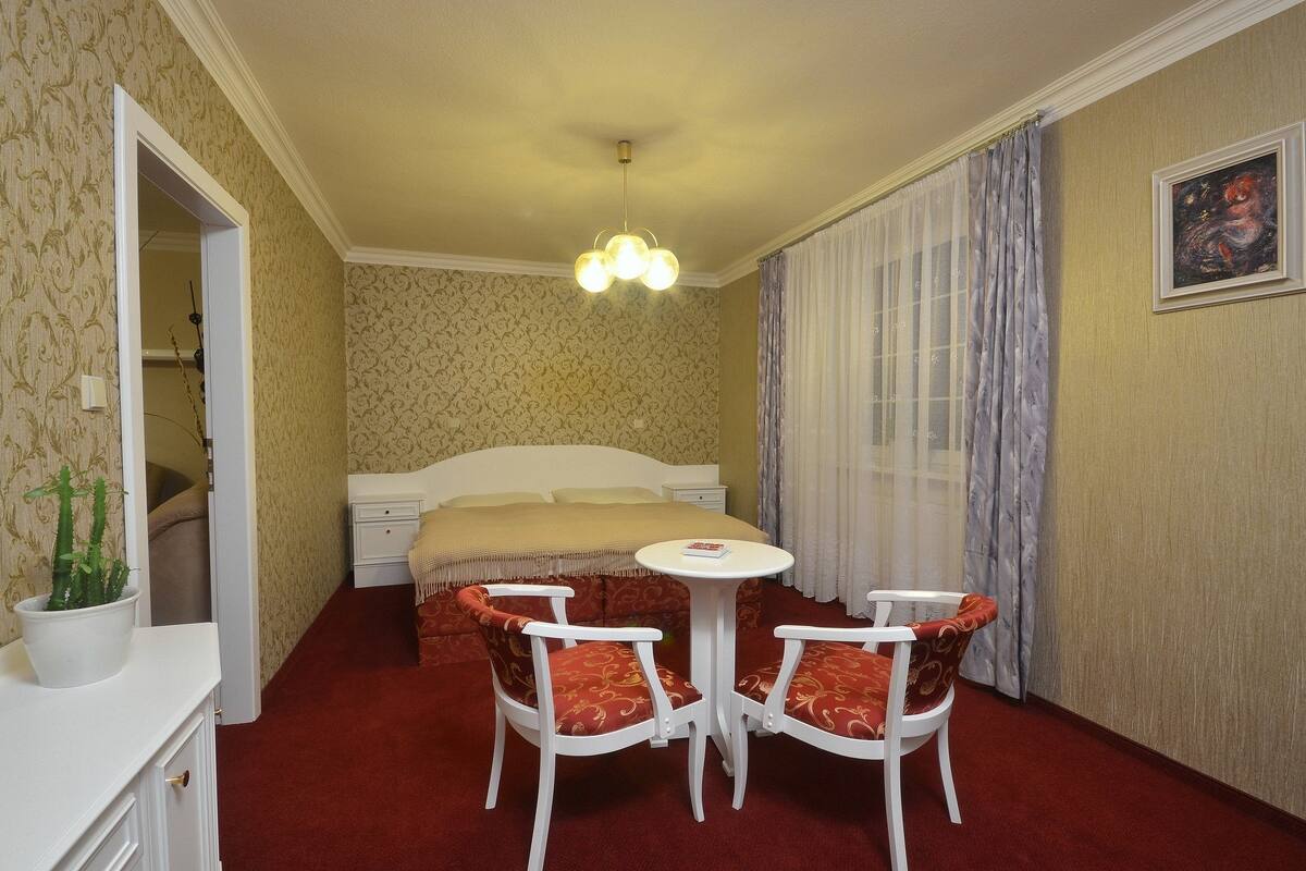 A hotel room with a bed, a small table with two chairs, a potted plant, and a painting on the wall.