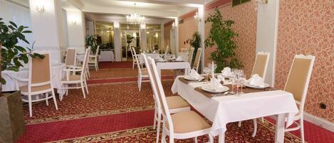 Un comedor formal con mesas y sillas blancas, piso con alfombra roja y una araña.