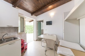 A compact kitchen with a sink, a dining area with a table and chairs, and a bed with a white mattress. - Nicolaus Club Alimini Smile  (Otranto)