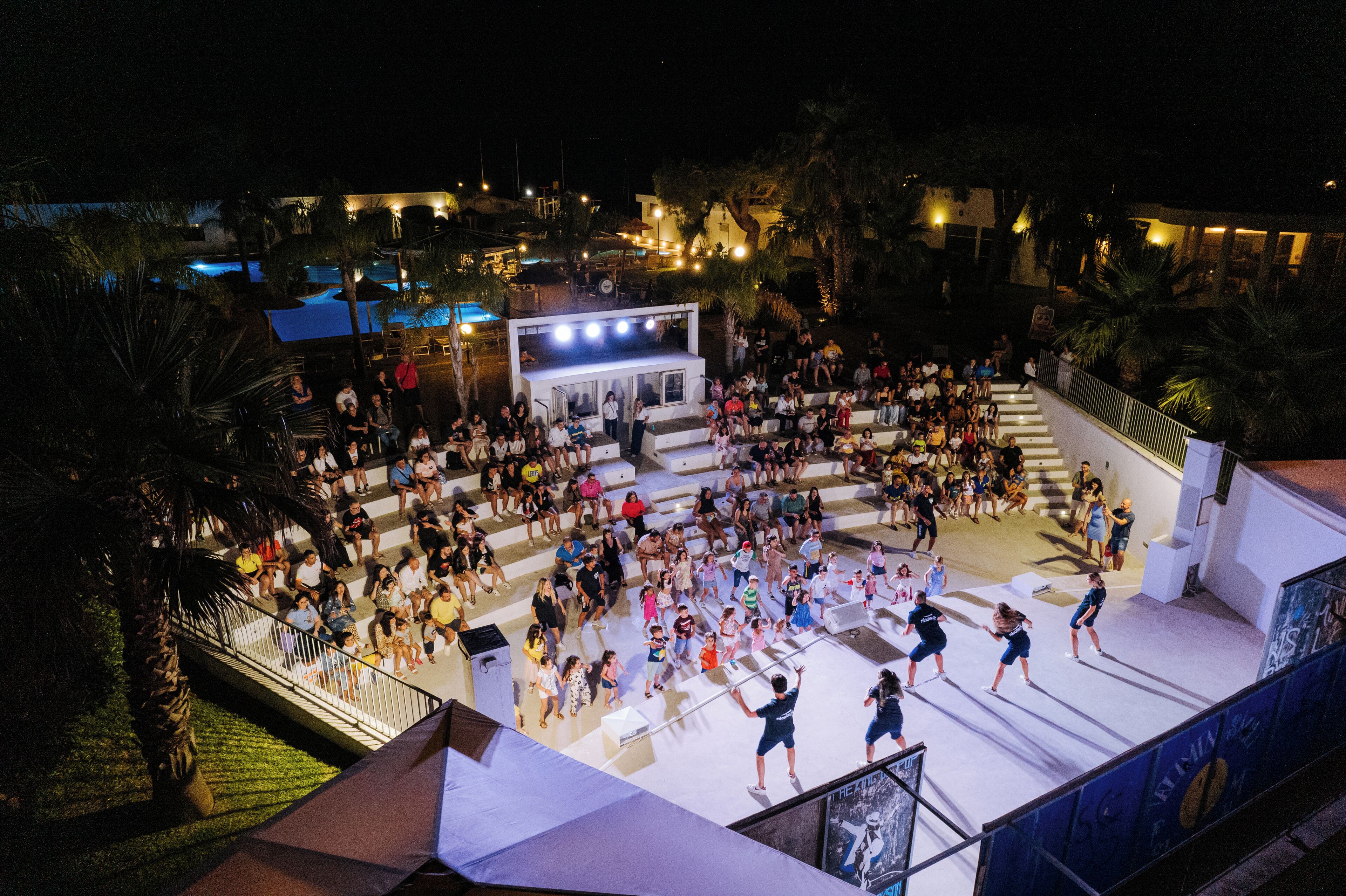 Ampia area esterna per eventi con un palco e posti a sedere, circondata da palme e edifici.