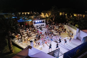 A large outdoor gathering area with a stage and seating, surrounded by palm trees and buildings. - Nicolaus Club Alimini Smile  (Otranto)