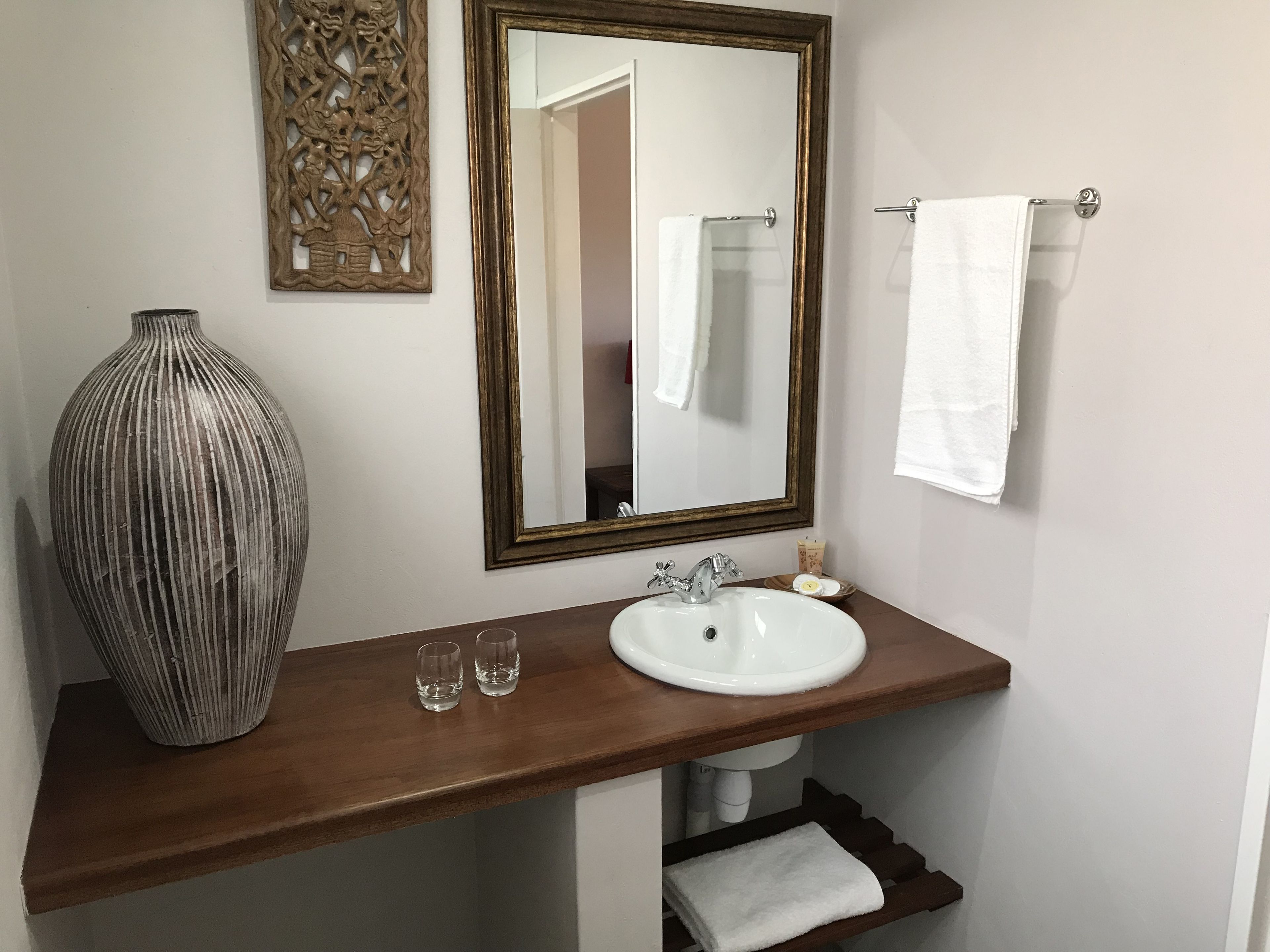 A bathroom with a wooden vanity, a white sink, two glasses, a large vase, a mirror, and a towel rack.