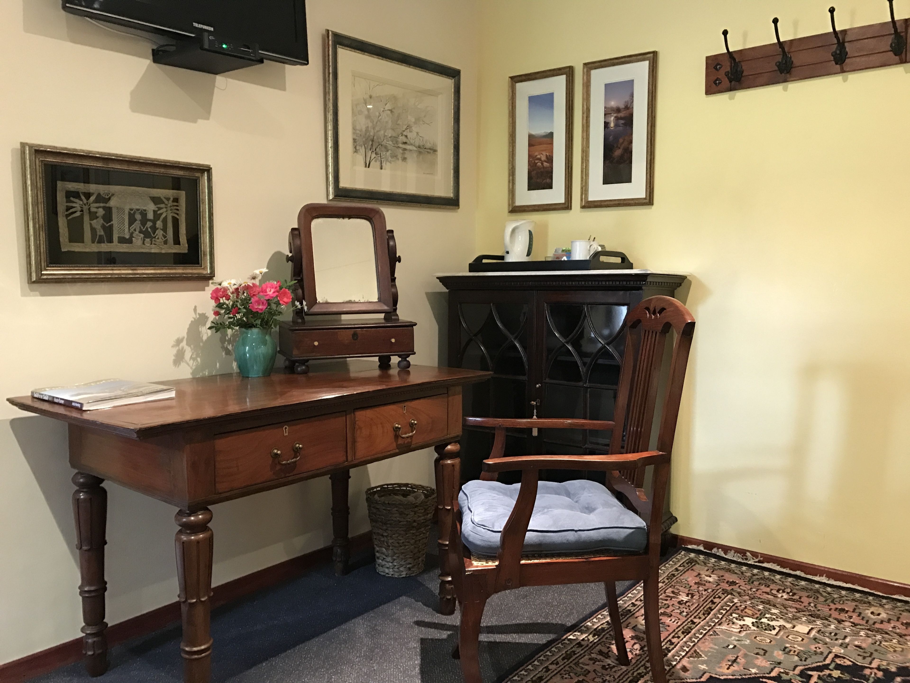 A wooden desk with drawers, a chair with a cushion, a mirror, and framed pictures on the wall.