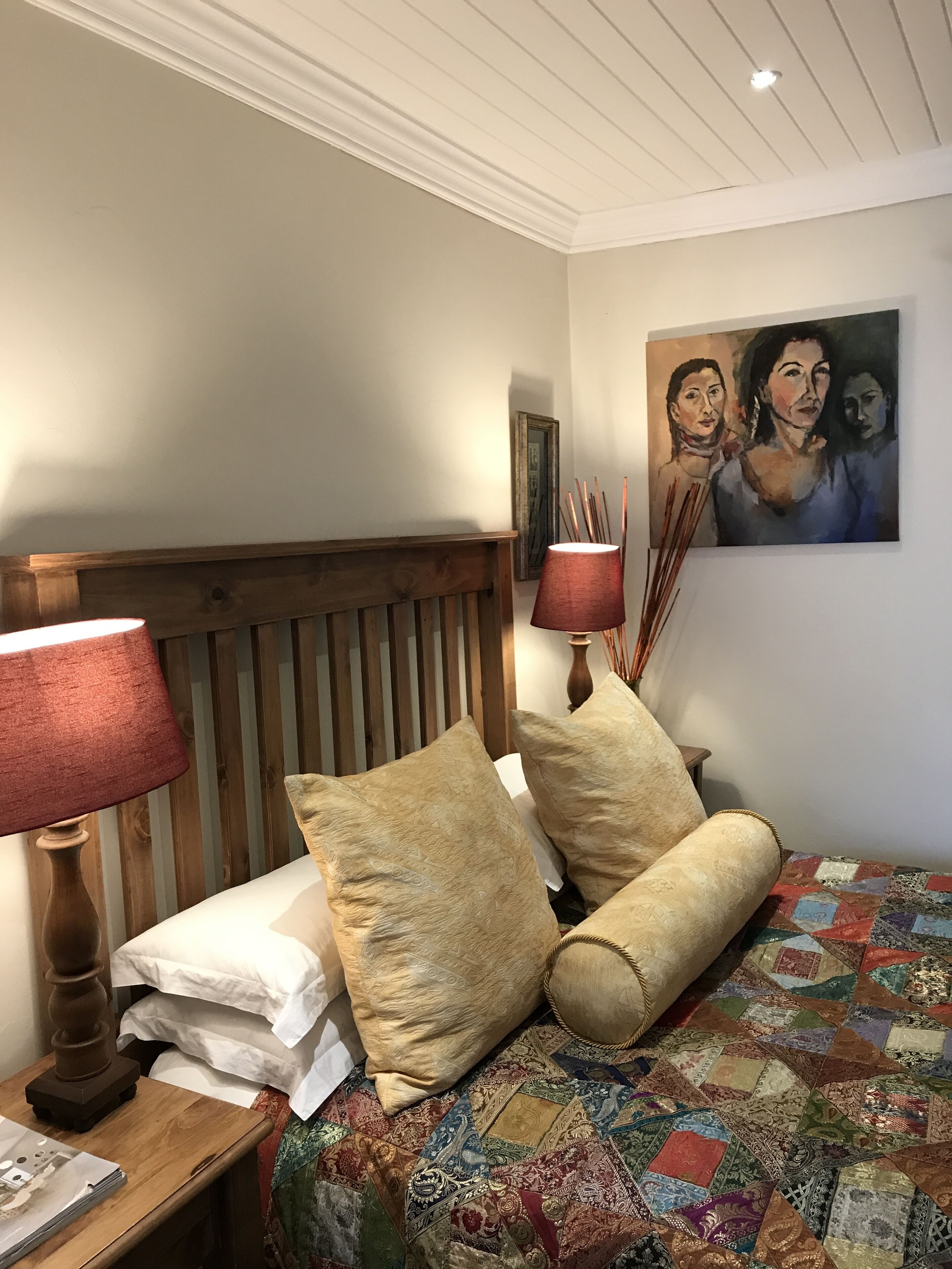 A bedroom with a wooden headboard, a wooden nightstand with a lamp, a quilted bedspread, and a painting on the wall.