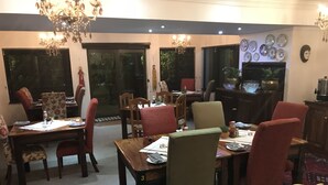A dining area with a large table, multiple chairs, and a chandelier. There are windows with blinds and a television on a stand. - The Crown Guesthouse (Bethlehem)