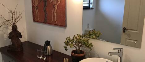 Un baño con encimera de madera, lavamanos blanco, planta en maceta, cafetera y un cuadro sobre el espejo.