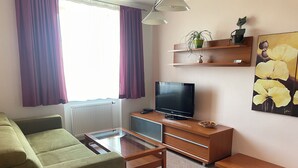 Una sala de estar moderna con un sofá verde, una mesa de centro de vidrio, un televisor de pantalla plana, estantes de madera con objetos decorativos y un cuadro de flores amarillas.