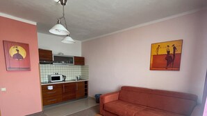 Una sala de estar con una mesa de centro de madera, un sofá rojo, una zona de cocina con armarios y un microondas, y dos cuadros en las paredes.
