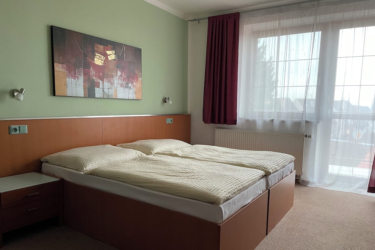Ein Hotelzimmer mit Doppelbett, Nachttisch, Lampe, einem Bild an der Wand und einem Fenster mit Vorhängen.