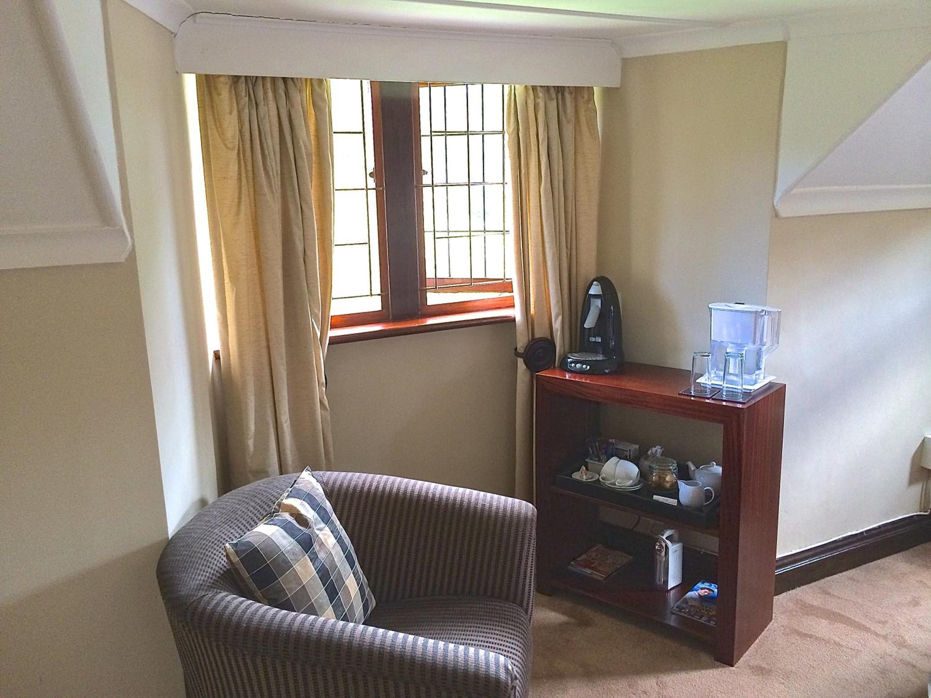 Una habitación de esquina con un sillón a rayas marrones, una mesita de madera con cafetera y dispensador de agua, y una ventana con cortinas color beige.