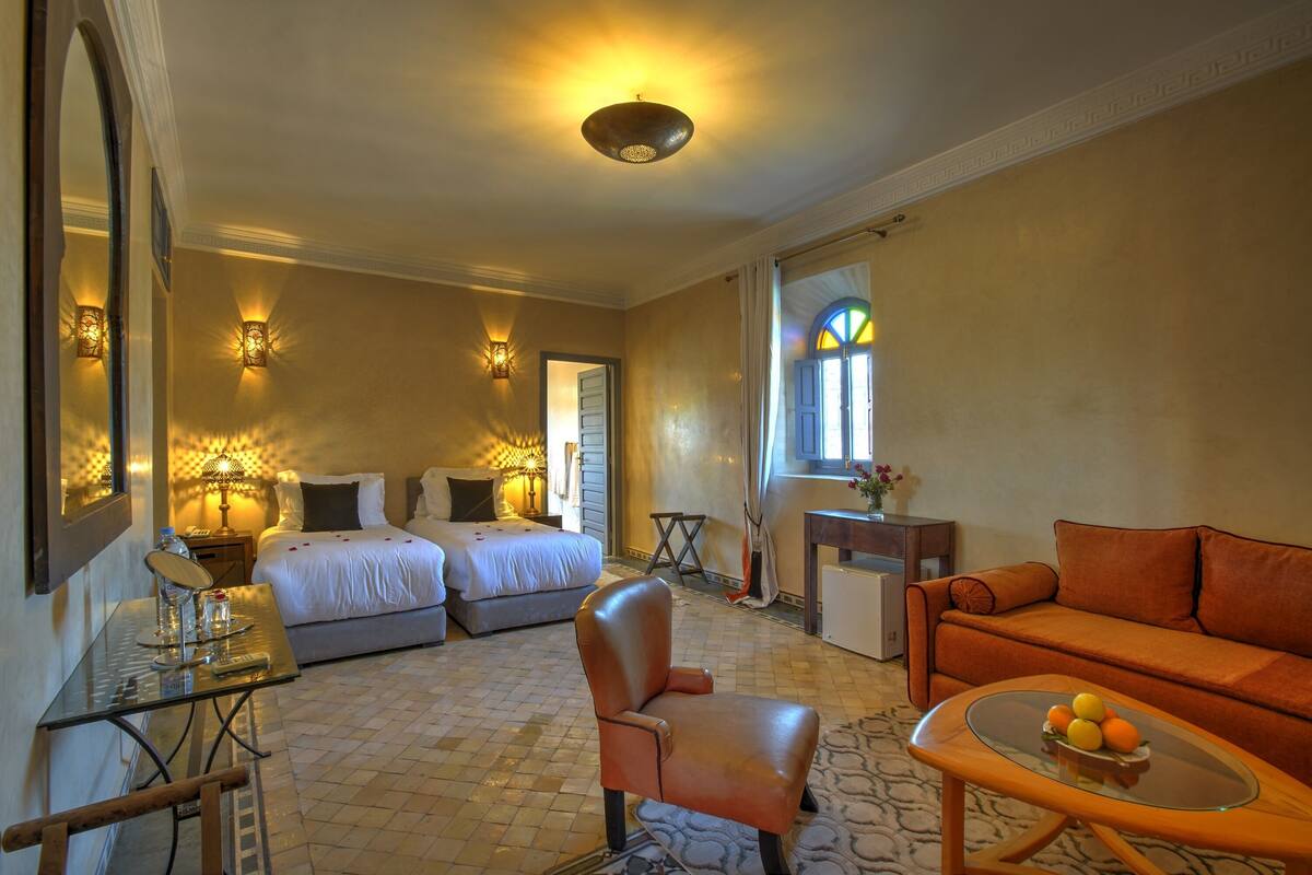 Quarto de hotel com duas camas, área de estar com sofá e poltrona, uma mesinha com frutas e um espelho.