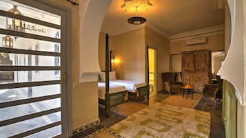 Habitación de hotel con cama, una zona de estar con sillón y mesa, una alfombra decorativa y vistas a través de un ventanal.