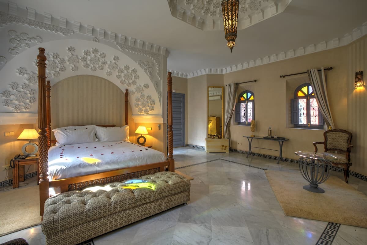 Um quarto espaçoso com uma cama de dossel, um espaço de estar com uma mesa e cadeiras, e um espelho decorativo.