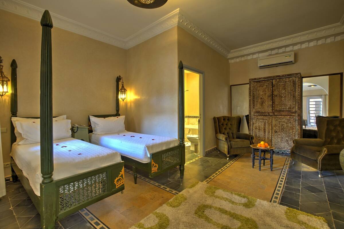 Quarto de hotel com duas camas, um espaço de estar com uma cadeira e uma pequena mesa, e um banheiro visível através de uma porta aberta.