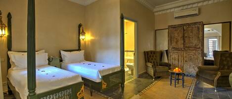 A hotel room with two beds, a sitting area with a chair and a small table, and a bathroom visible through an open door.