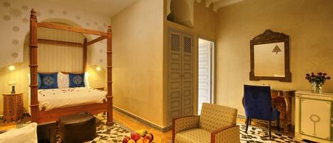A four-poster bed with a fruit bowl on the side, a patterned armchair, and a mirror on the wall.