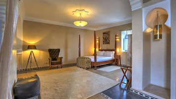A bedroom with a four-poster bed, a sitting area with a chair and ottoman, a large rug, and a chandelier.