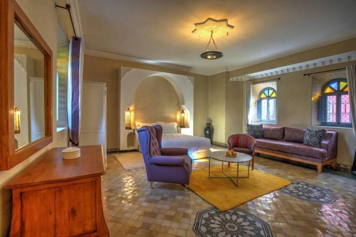 Quarto de hotel com cama, poltrona roxa, sofá, mesa redonda pequena e cómoda de madeira.