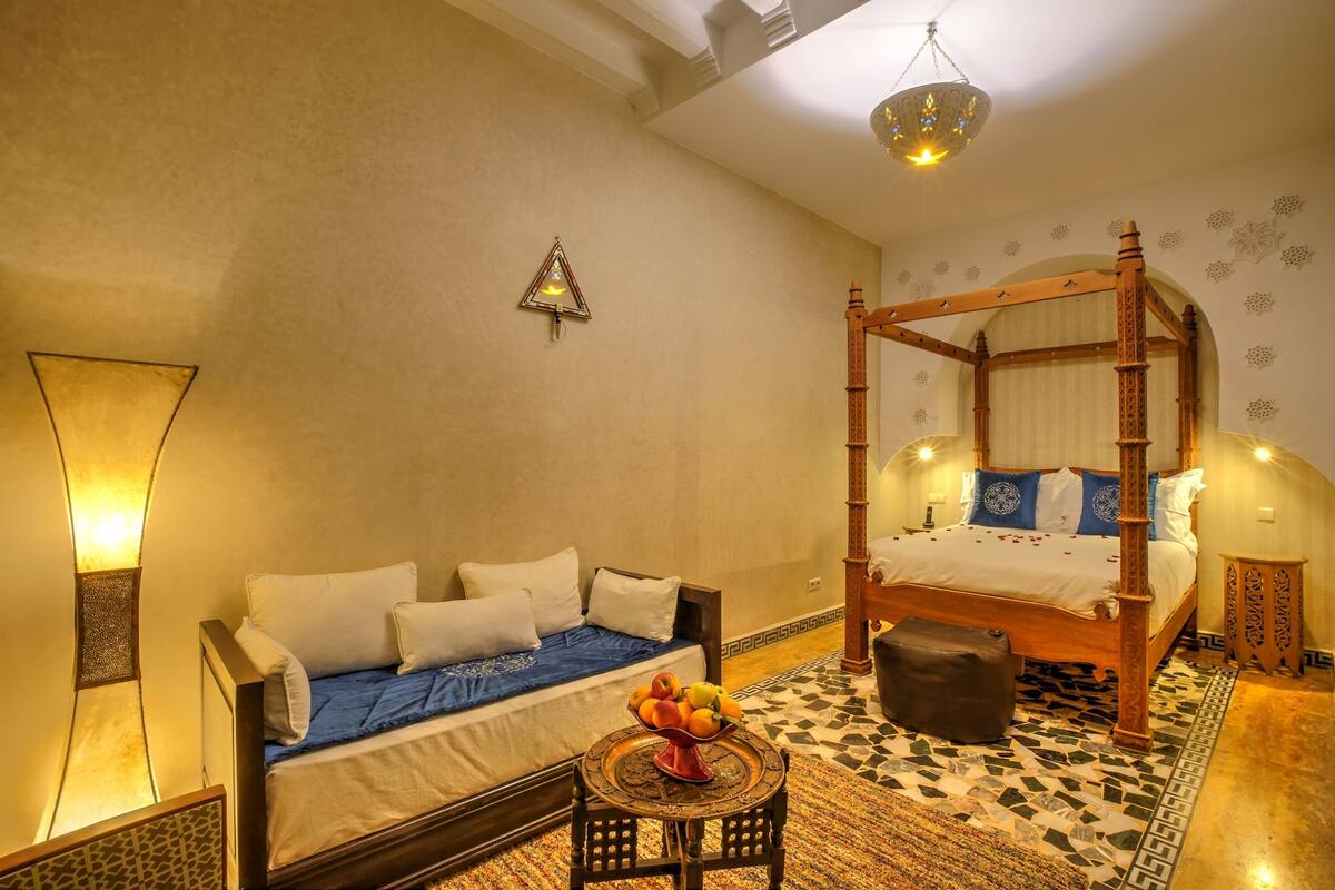 Quarto com cama de dossel, sofá, mesinha com frutas e um objeto decorativo na parede.