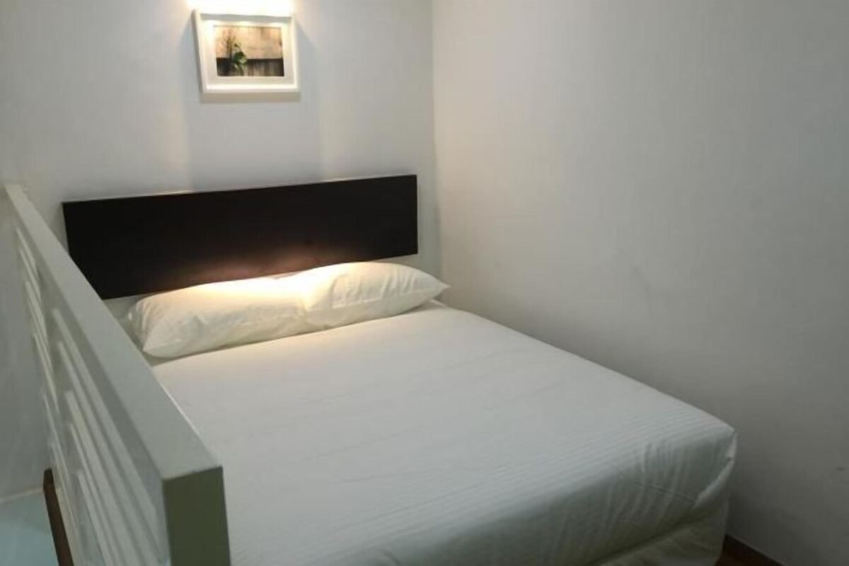 Una cama pequeña y bien hecha, con un colchón y almohadas blancos, un cabecero oscuro y un cuadro enmarcado en la pared superior.
