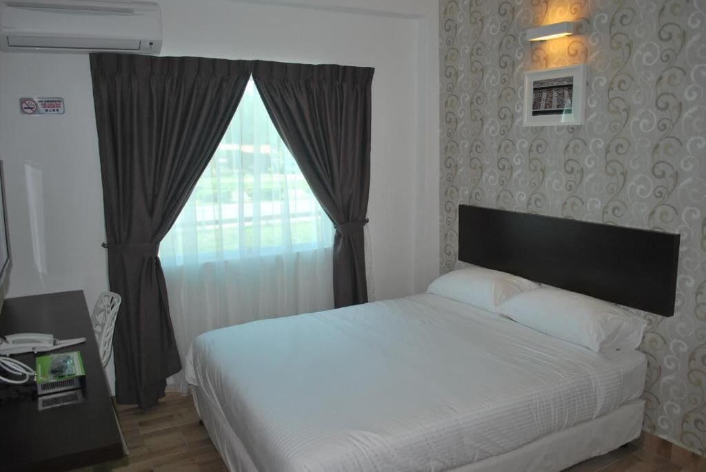 A hotel room with a bed, a desk with a telephone and a small electronic device, a window with curtains, and a wall-mounted light fixture.