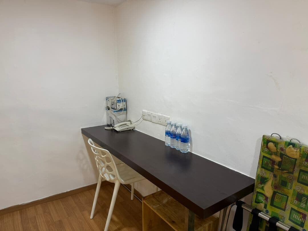 A small, simple room with a dark wooden desk, a white chair, a telephone, and several water bottles.