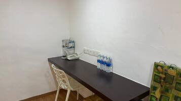 A small, simple room with a dark wooden desk, a white chair, a telephone, and several water bottles.