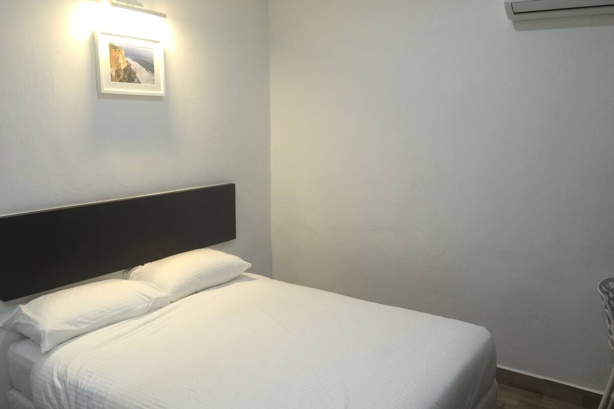 A neatly made bed with white linens in a room with a plain white wall and a small picture frame.