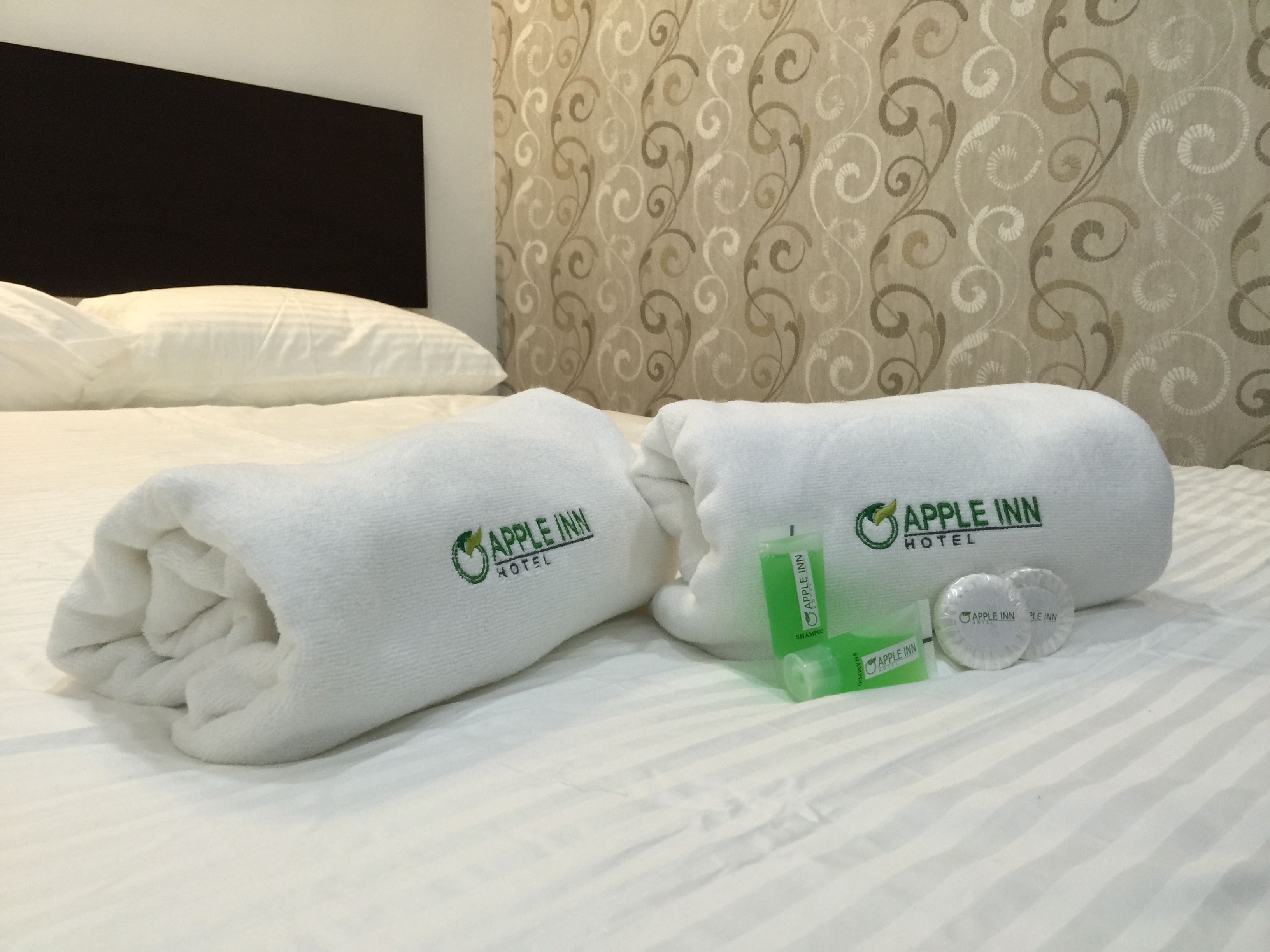 A neatly made bed with two towels and a bottle of lotion, both branded with the Apple Inn Hotel logo.