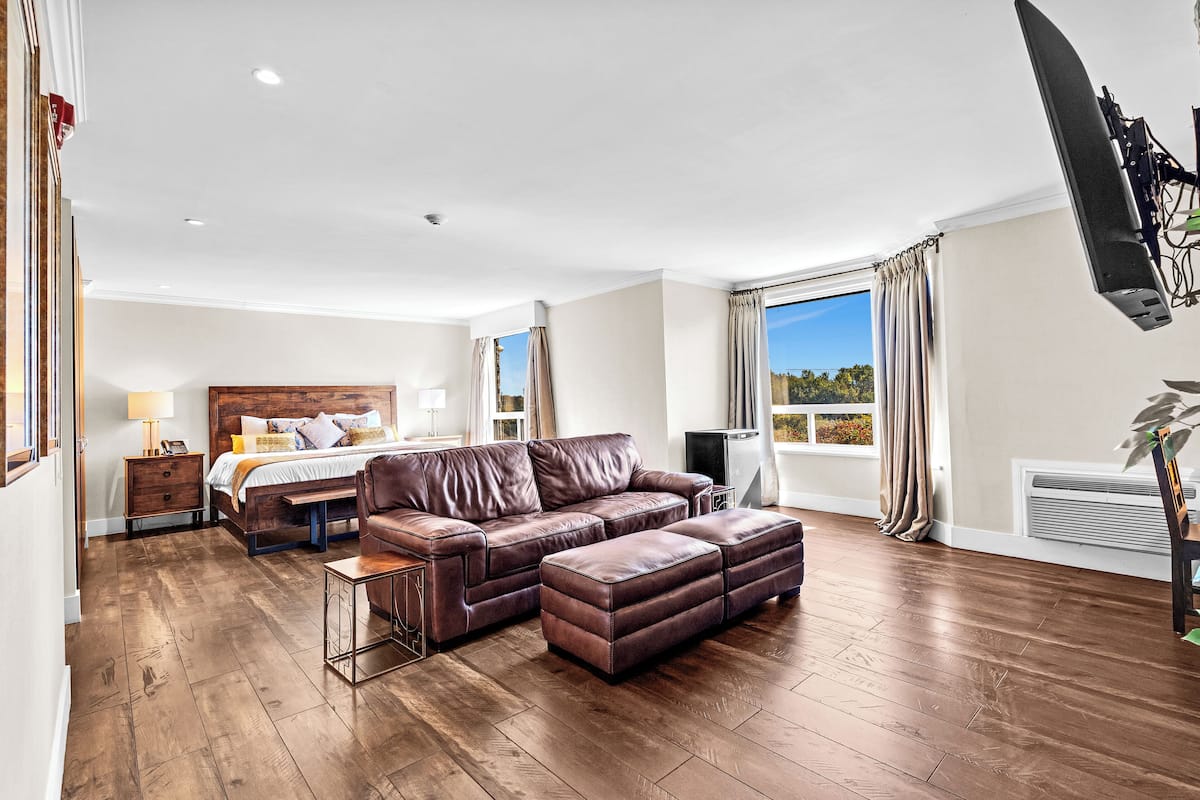 A spacious room with a large leather sofa, a bed with a wooden headboard, and a large window with curtains.