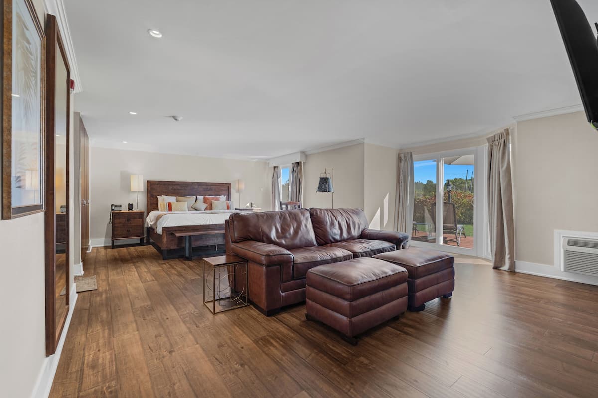 A spacious living room with a brown leather sofa and ottoman, a wooden floor, and a large window offering a view of the outdoors.