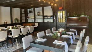 Ein Essbereich mit einem langen Tisch, der für eine Mahlzeit gedeckt ist, umgeben von Stühlen. Es gibt eine Bar mit Flaschen und Gläsern, und im Hintergrund steht ein Klavier.
