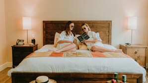 Ein Schlafzimmer mit zwei Personen, die auf dem Bett sitzen. Ein Nachttisch mit einer Lampe, einem Telefon und einer Tasse steht daneben. Auf einem Tablett auf dem Boden liegen ein Hut und Äpfel.