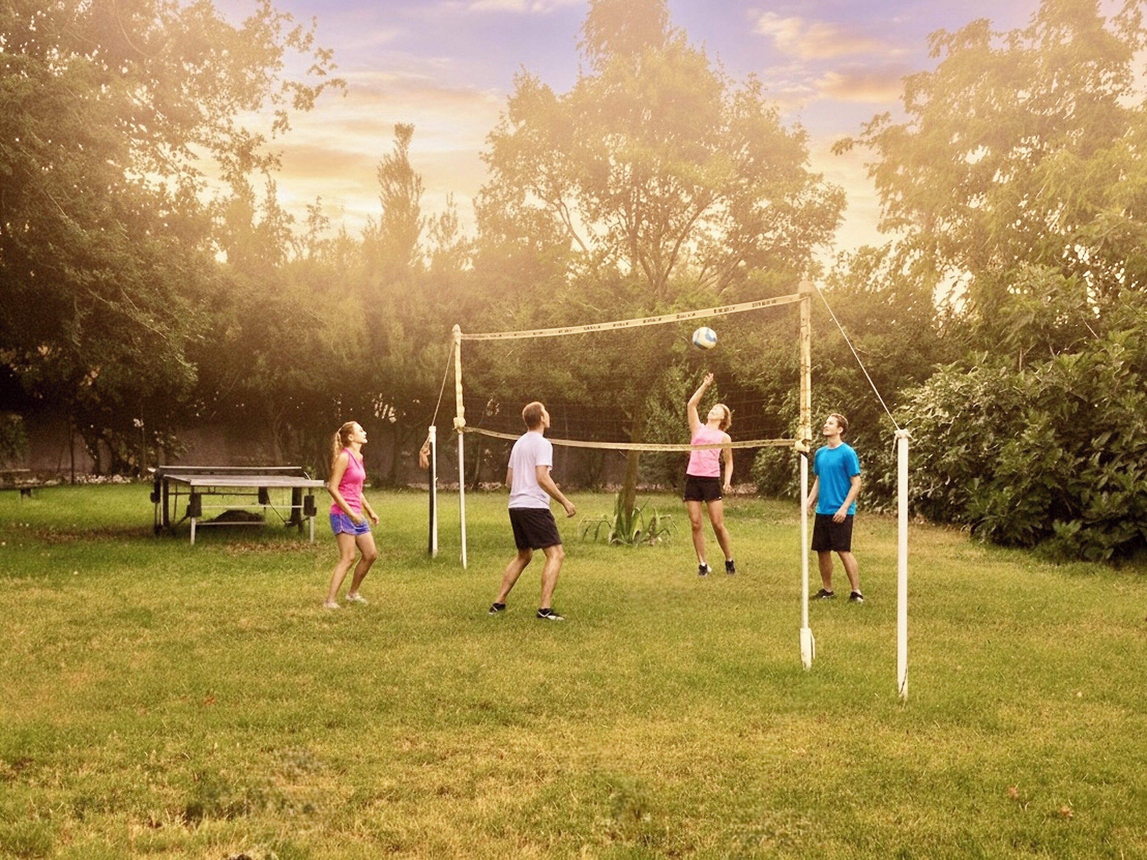 A group of people playing volleyball in a grassy outdoor area with trees in the background.