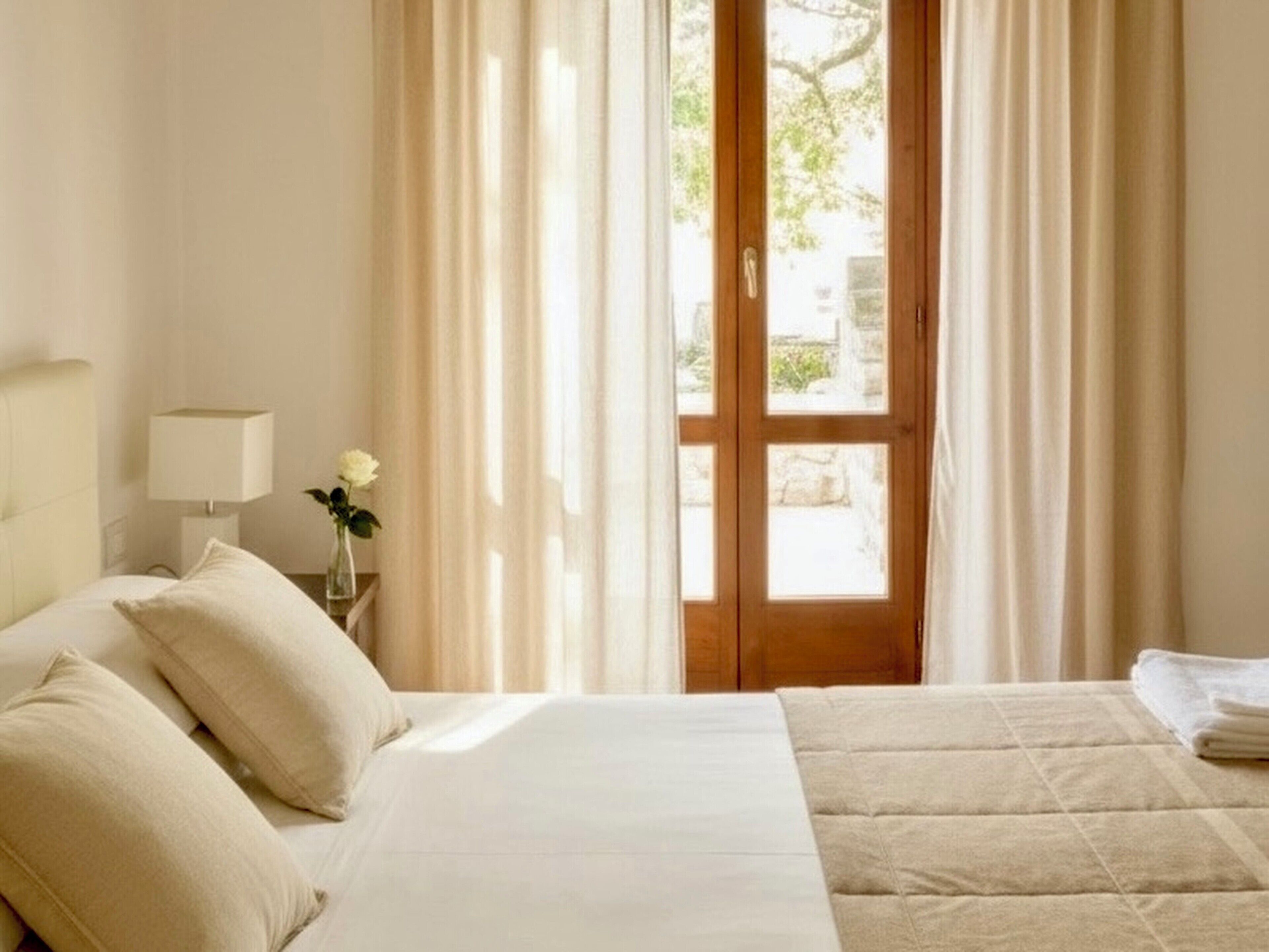 Una camera da letto con un letto, un comodino con una lampada, un vaso con un fiore e vista attraverso una finestra con tende.