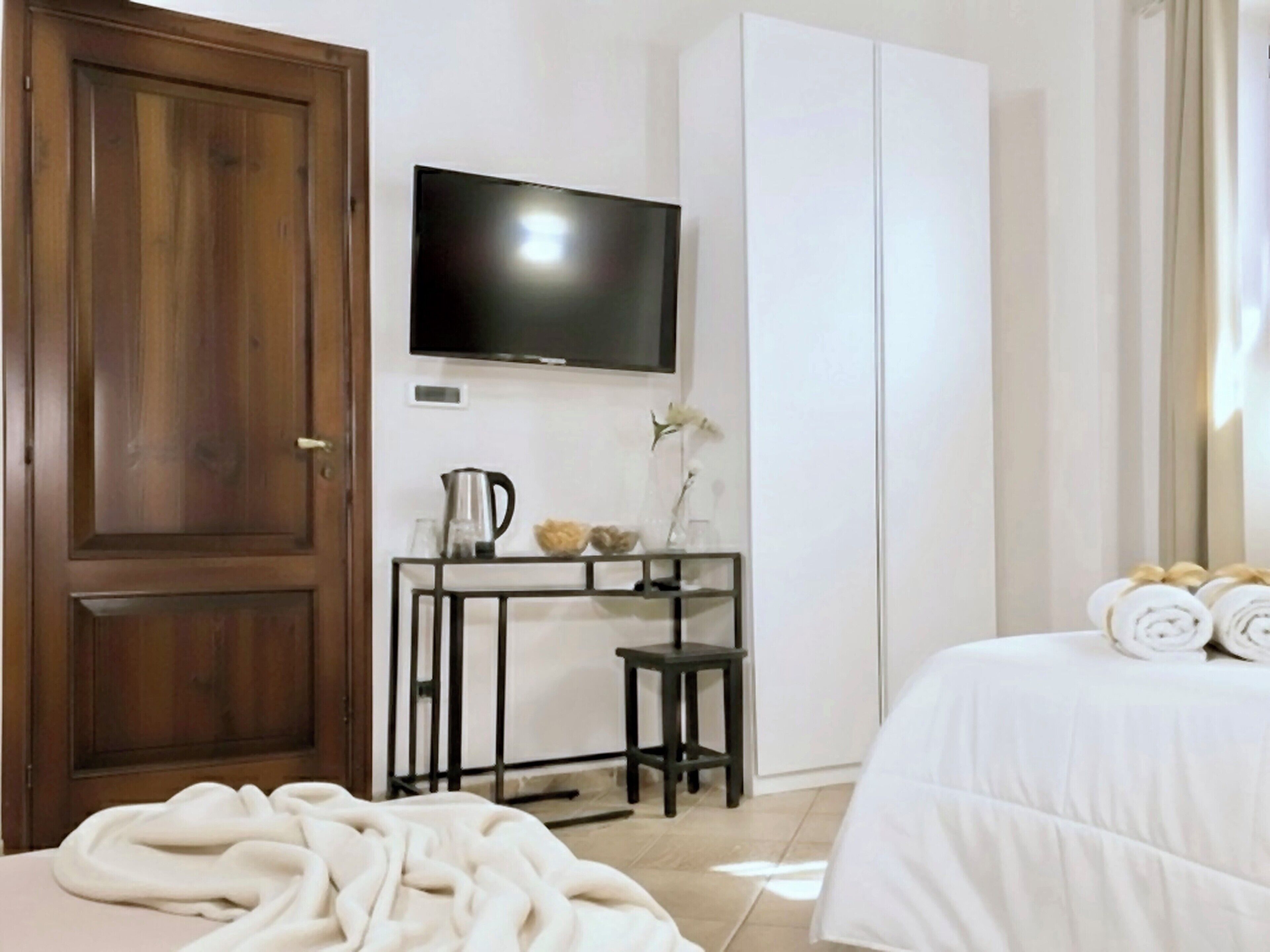 Une chambre d’hôtel avec une porte en bois, un téléviseur à écran plat fixé au mur, une petite table avec une bouilloire et des objets décoratifs, un lit avec des serviettes pliées et une grande armoire.