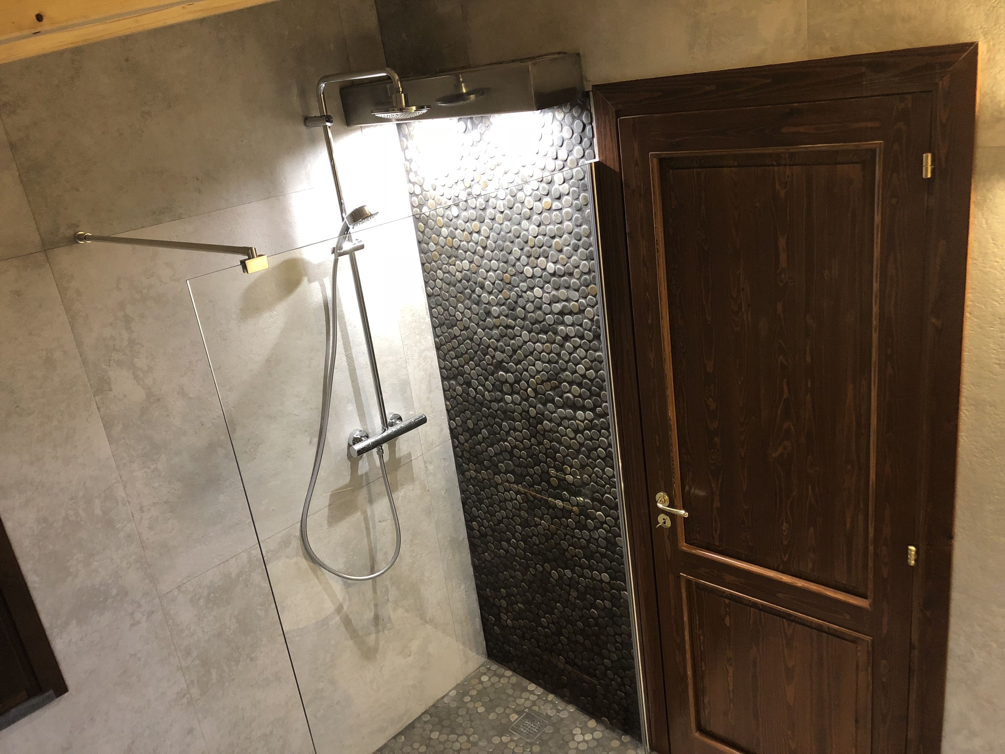 Una doccia con soffione a mano e rubinetto a parete, una porta in legno e un muro dalla superficie strutturata.