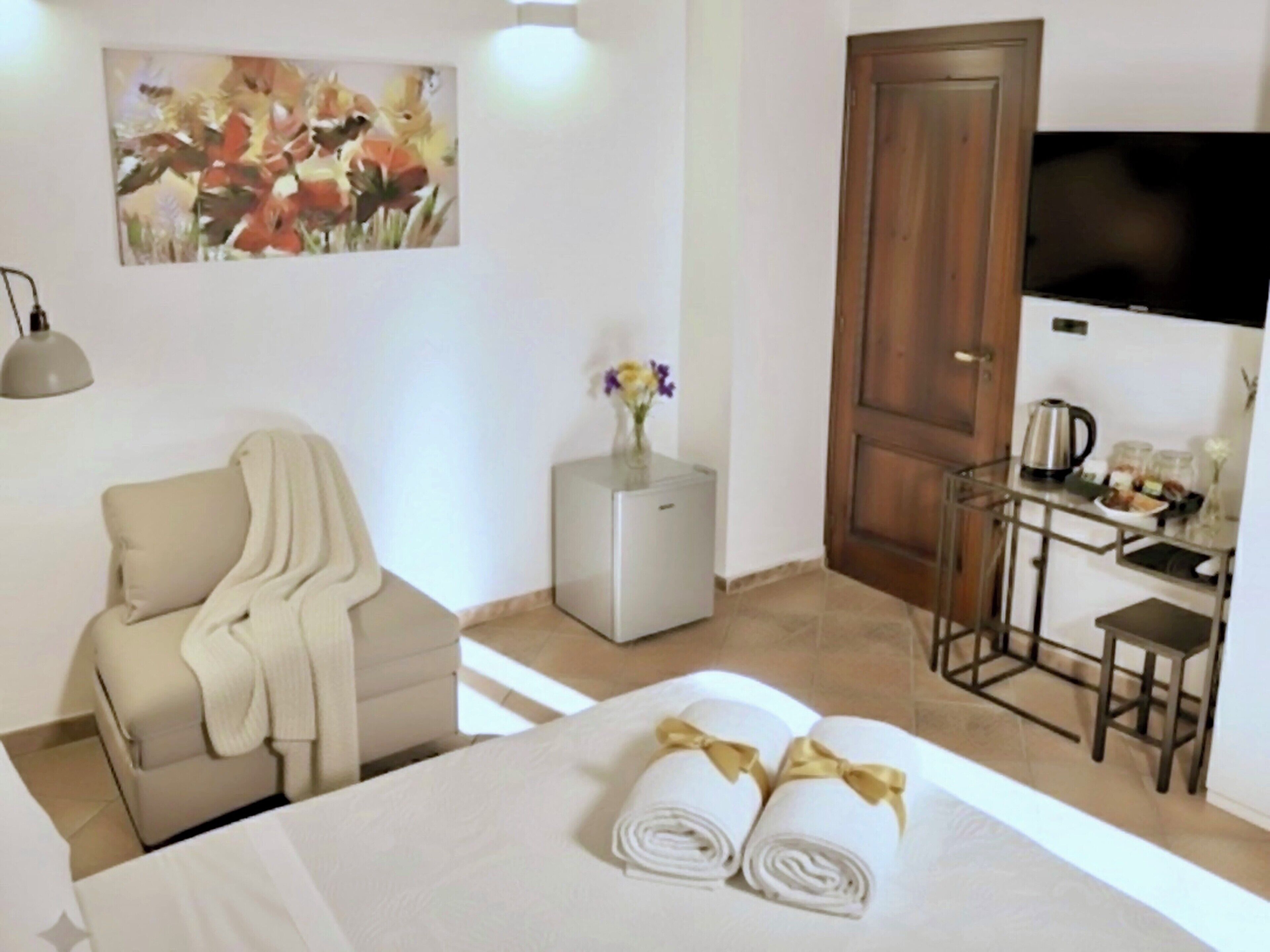 Quarto de hotel com uma cama, uma poltrona, uma mesinha com um vaso de flores, televisão e uma área de kitchenette.