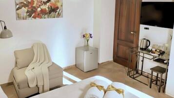 Quarto de hotel com uma cama, uma poltrona, uma mesinha com um vaso de flores, televisão e uma área de kitchenette.