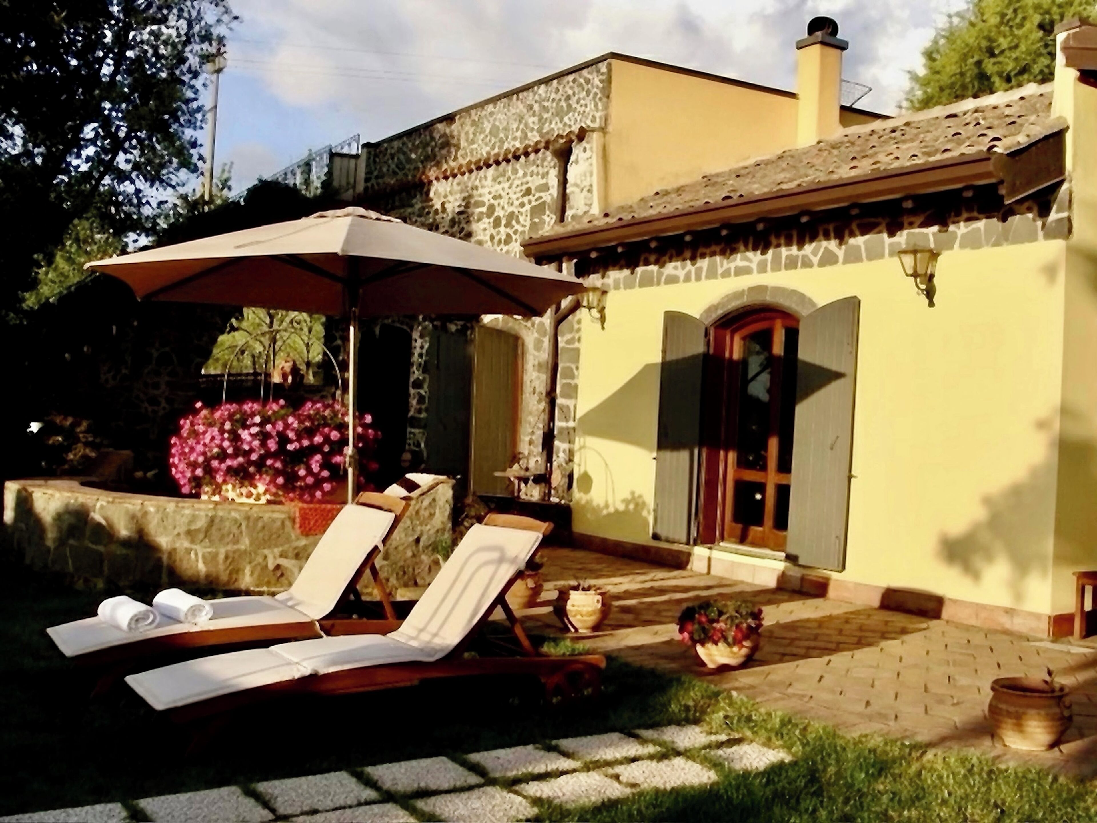 A patio with lounge chairs and a pergola, a stone wall with flowers, and a building with a tiled roof and a wooden door.