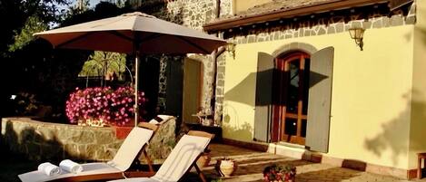 A patio with lounge chairs and a pergola, a stone wall with flowers, and a building with a tiled roof and a wooden door.