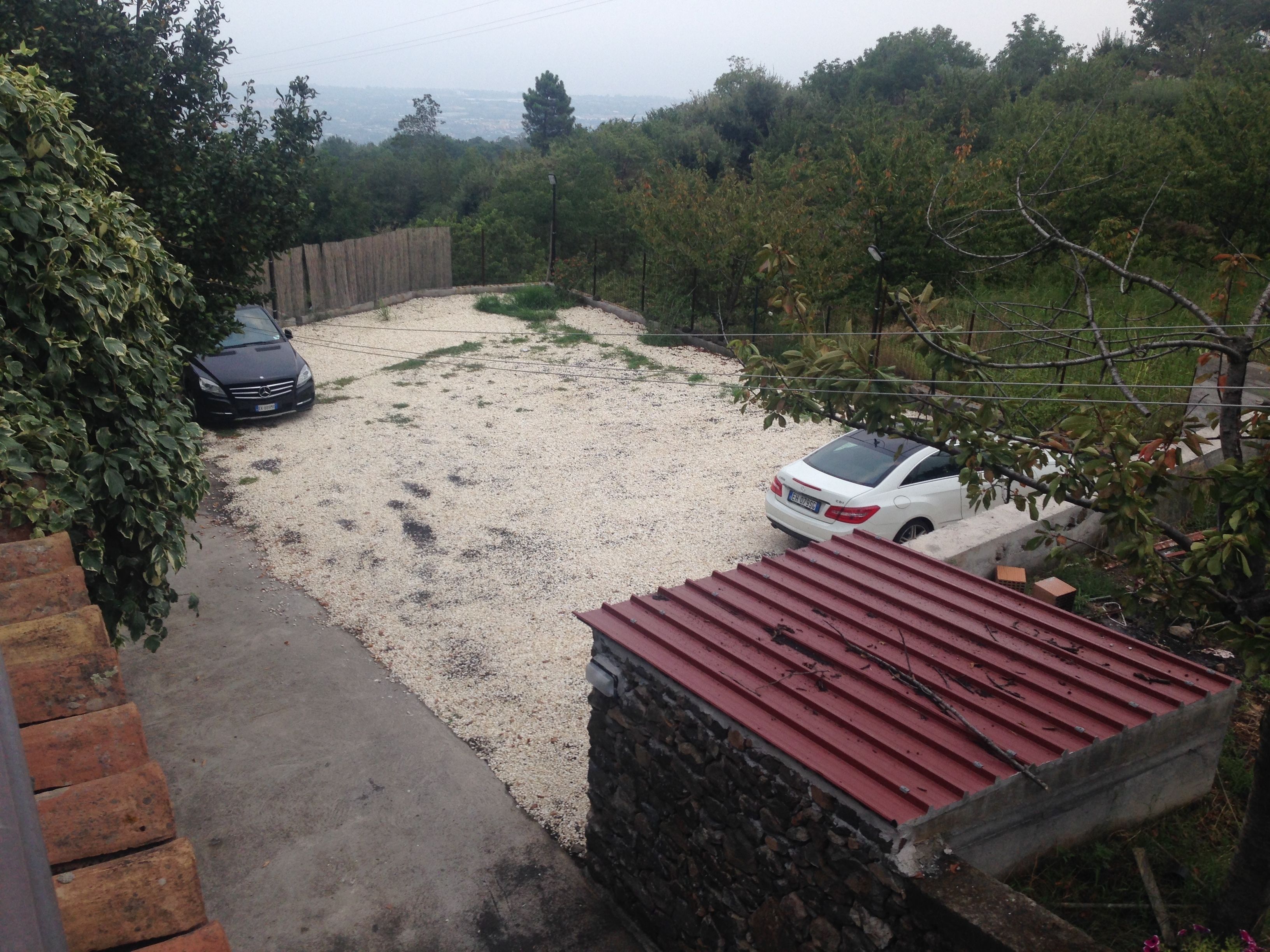 A gravel driveway leading to a white car parked on a gravel area, with a red-roofed structure on the right and a black car on the left.